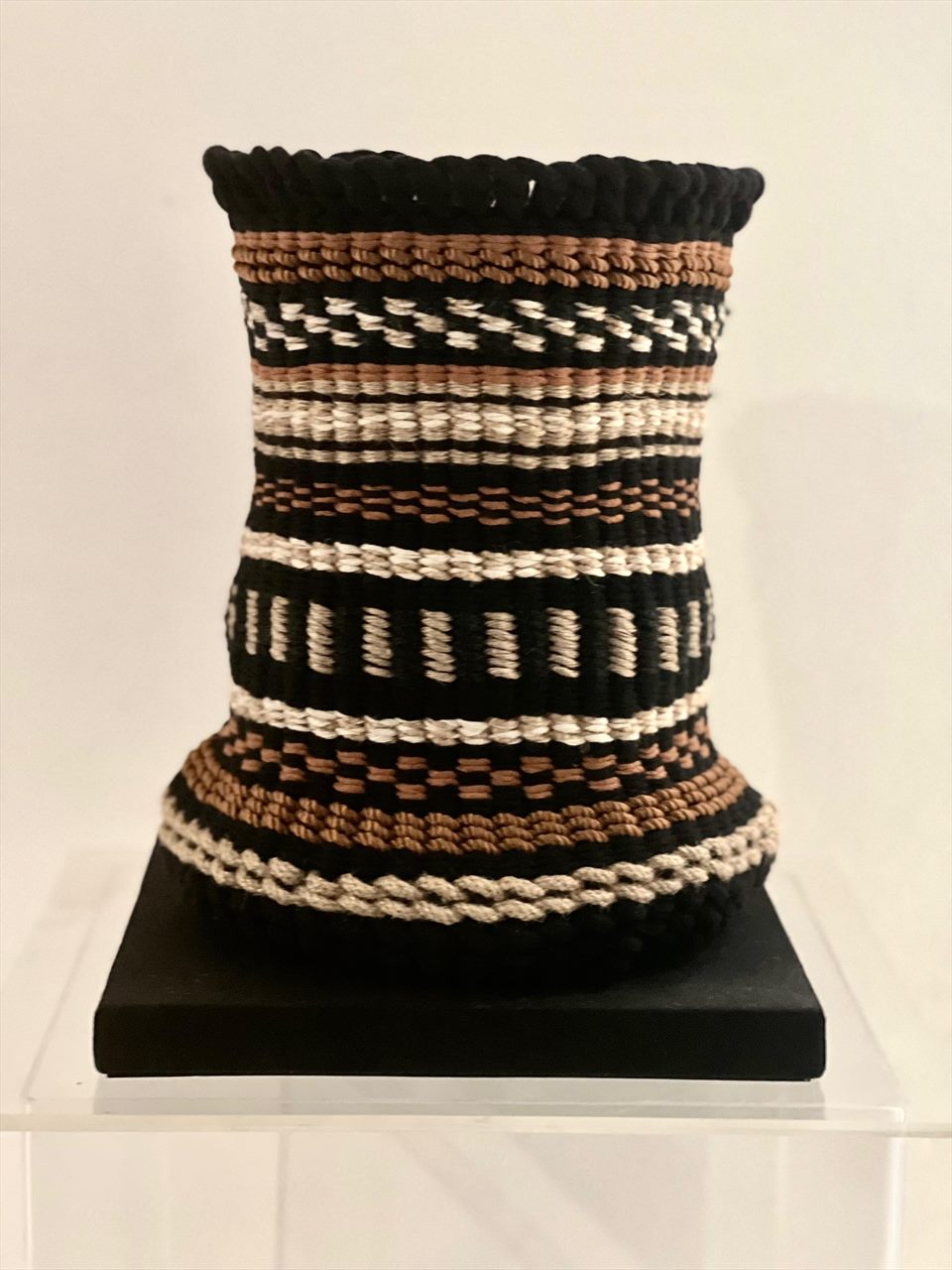 Vasija de cestería de talle estrecho tejida en negro, cobre y crudo, sobre base negra en sala de exposición.