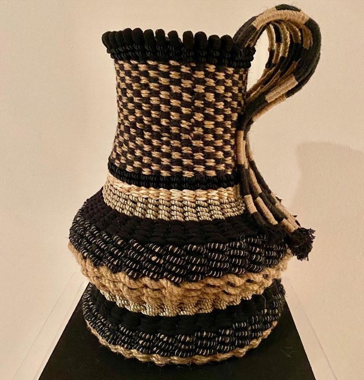 Cántaro tejido en negro y fibra cruda, con asa trenzada y cuerpo de damero.