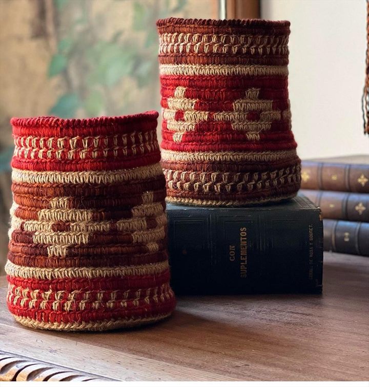 Dos cestos cilíndricos tejidos en rojo, tierra y crudo con grecas, apoyados sobre libros antiguos.