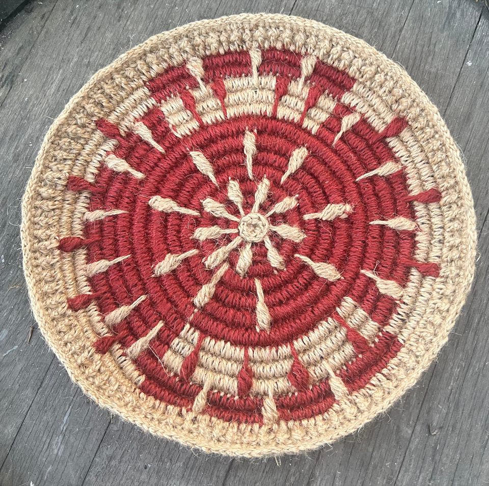 Pieza circular de cestería tejida en fibra natural: rayos crudos sobre campo rojo, apoyada sobre madera envejecida.