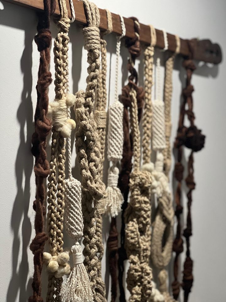 Serie de trenzados y cordones de fibra natural colgando de una vara de madera, vistos en perspectiva contra un muro claro.