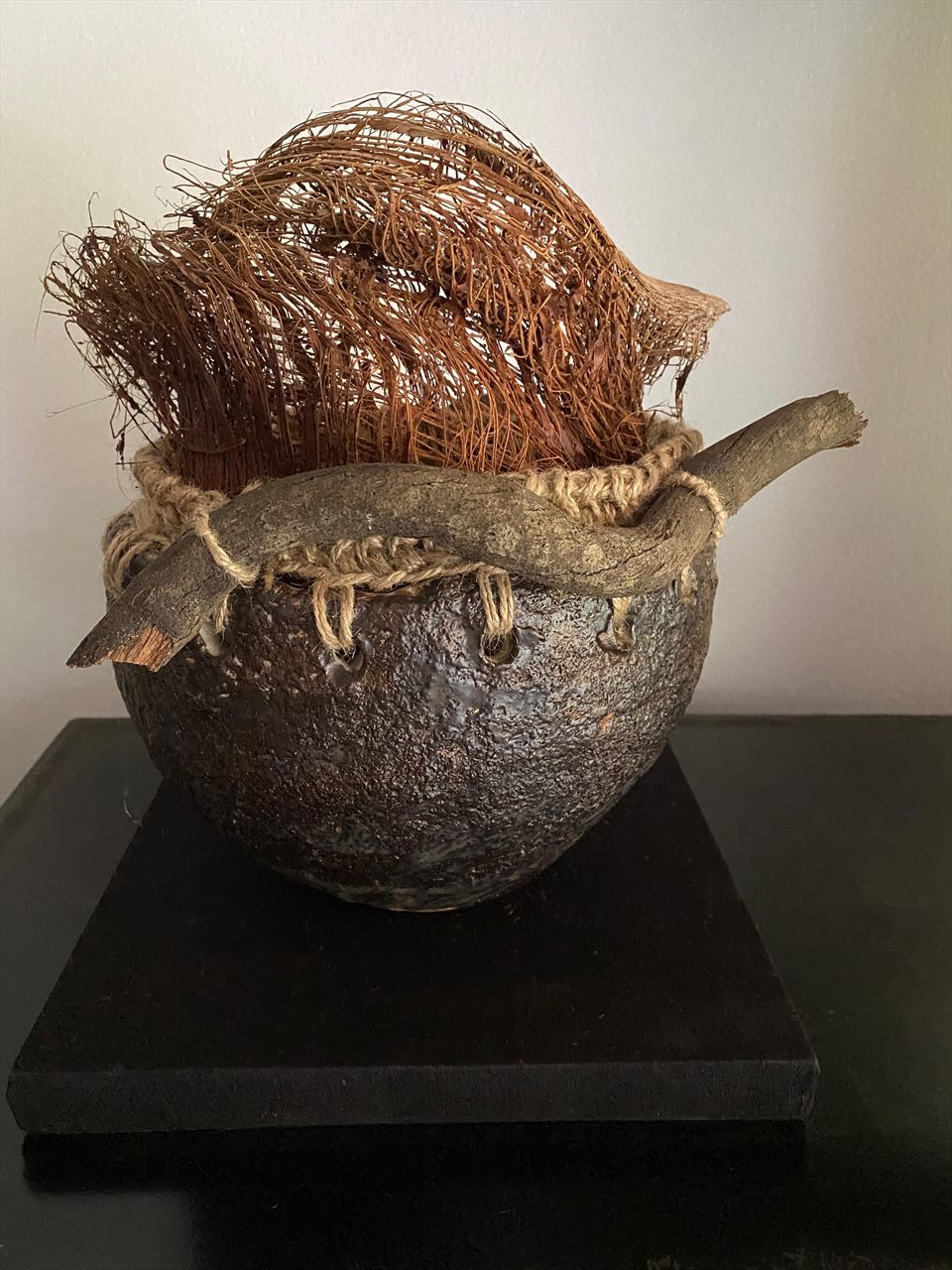 Huaco de piedra rugosa amarrado con cuerda de fibra, del que emerge un penacho de filamentos cobrizos.