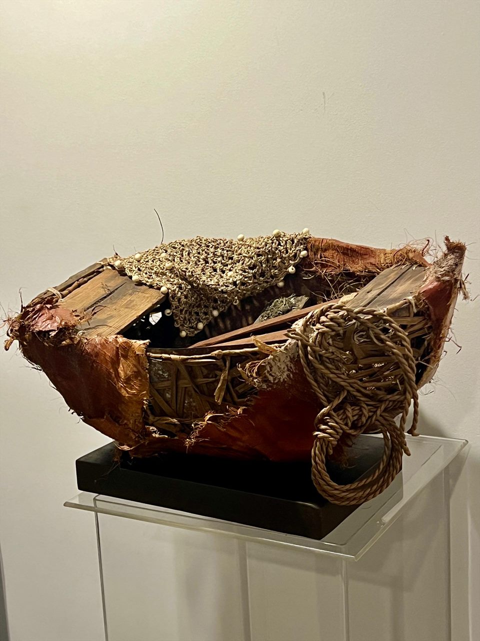 Embarcación escultórica tejida en fibra de coco y cuerda, con una vela de red anudada, sobre base negra en sala de exposición.