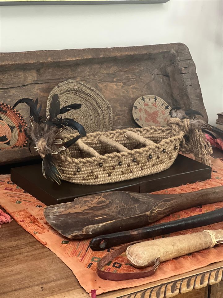 Embarcación tejida en cuerda con plumas negras en la proa, apoyada sobre una artesa de madera tallada junto a piezas de cestería.