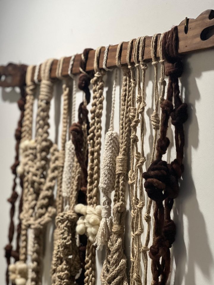 Quipu de fibras crudas y lana marrón colgado de una vara de madera antigua contra un muro blanco.