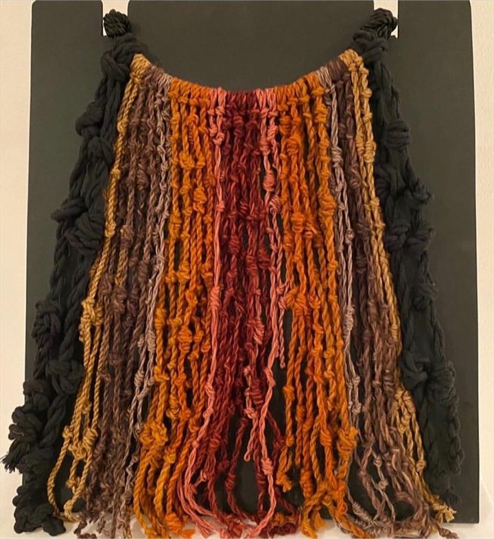Quipu de lanas gruesas en negro, ocre y rojo dispuesto sobre un panel oscuro.