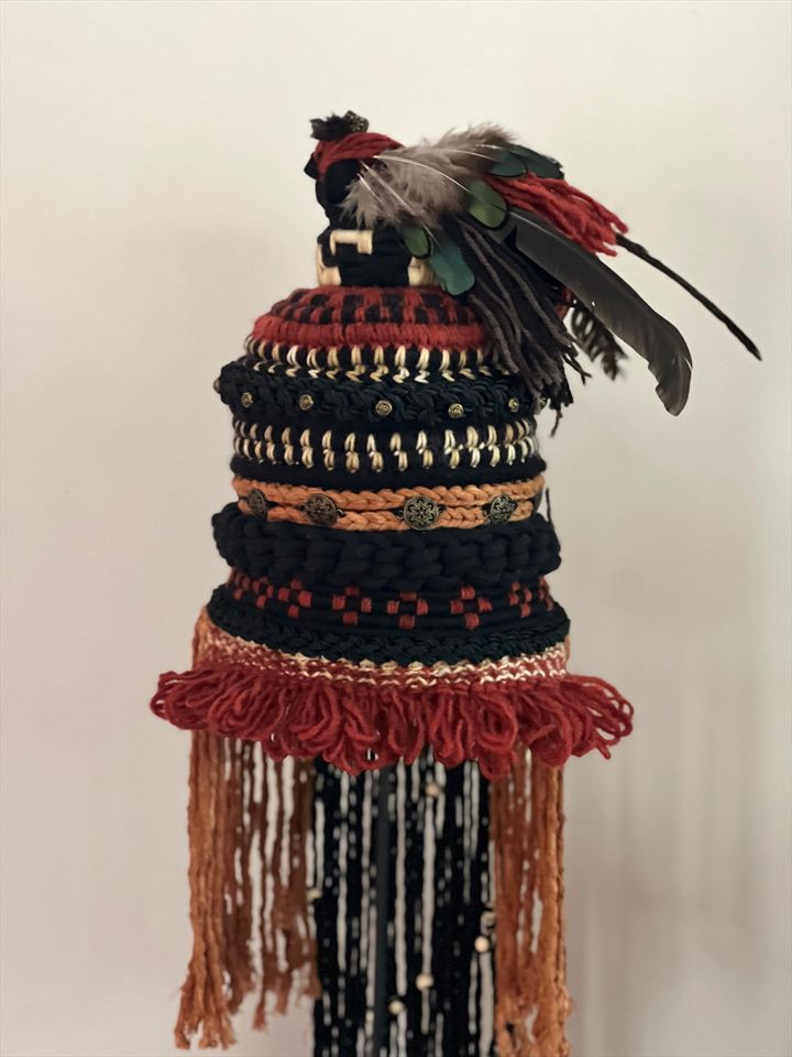 Tocado ceremonial tejido en rojo, negro y ocre, rematado con plumas y flecos largos, montado sobre pedestal.